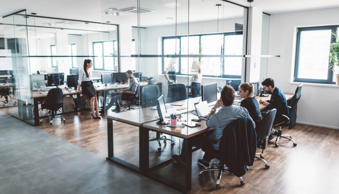 an open office space where 3 people are seated at their desks and another is standing leaning on a desk in the background.